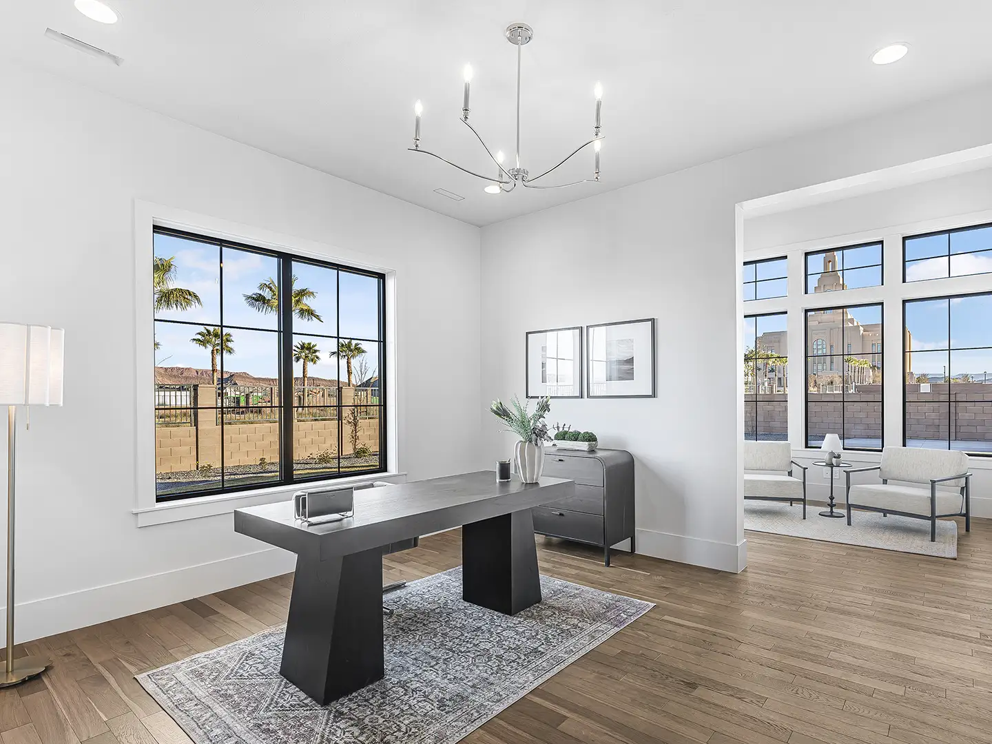 Modern home office with hardwood floors, dark wood desk, chandelier, and large windows with palm tree and temple views at Cedar Pointe.