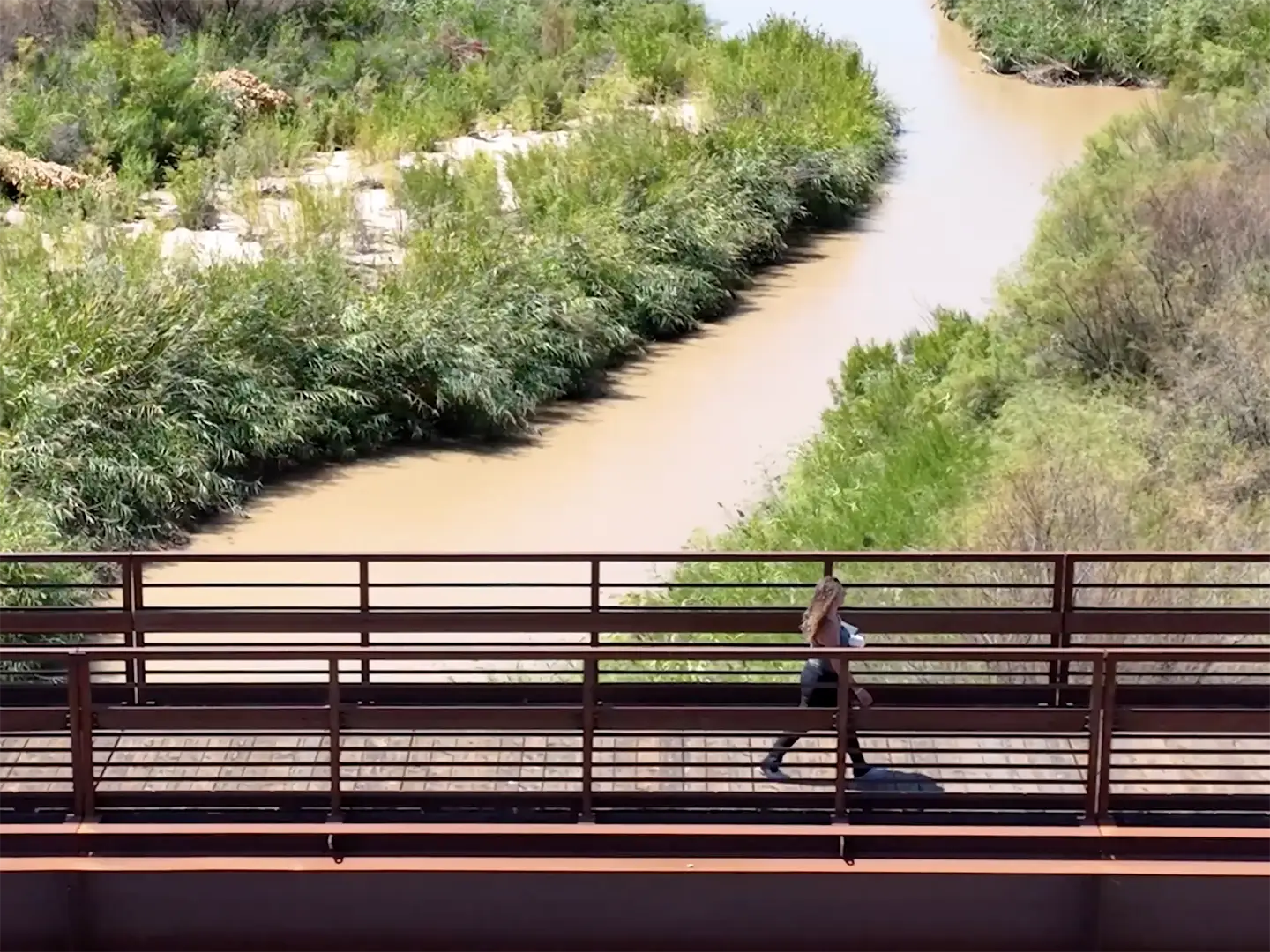Person walking across a metal pedestrian bridge over a winding dirt trail lined with desert greenery near St. George, Utah.