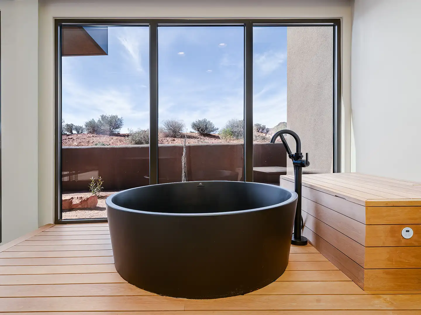Round black soaking tub on a raised wood platform with floor-to-ceiling glass and desert landscape views.
