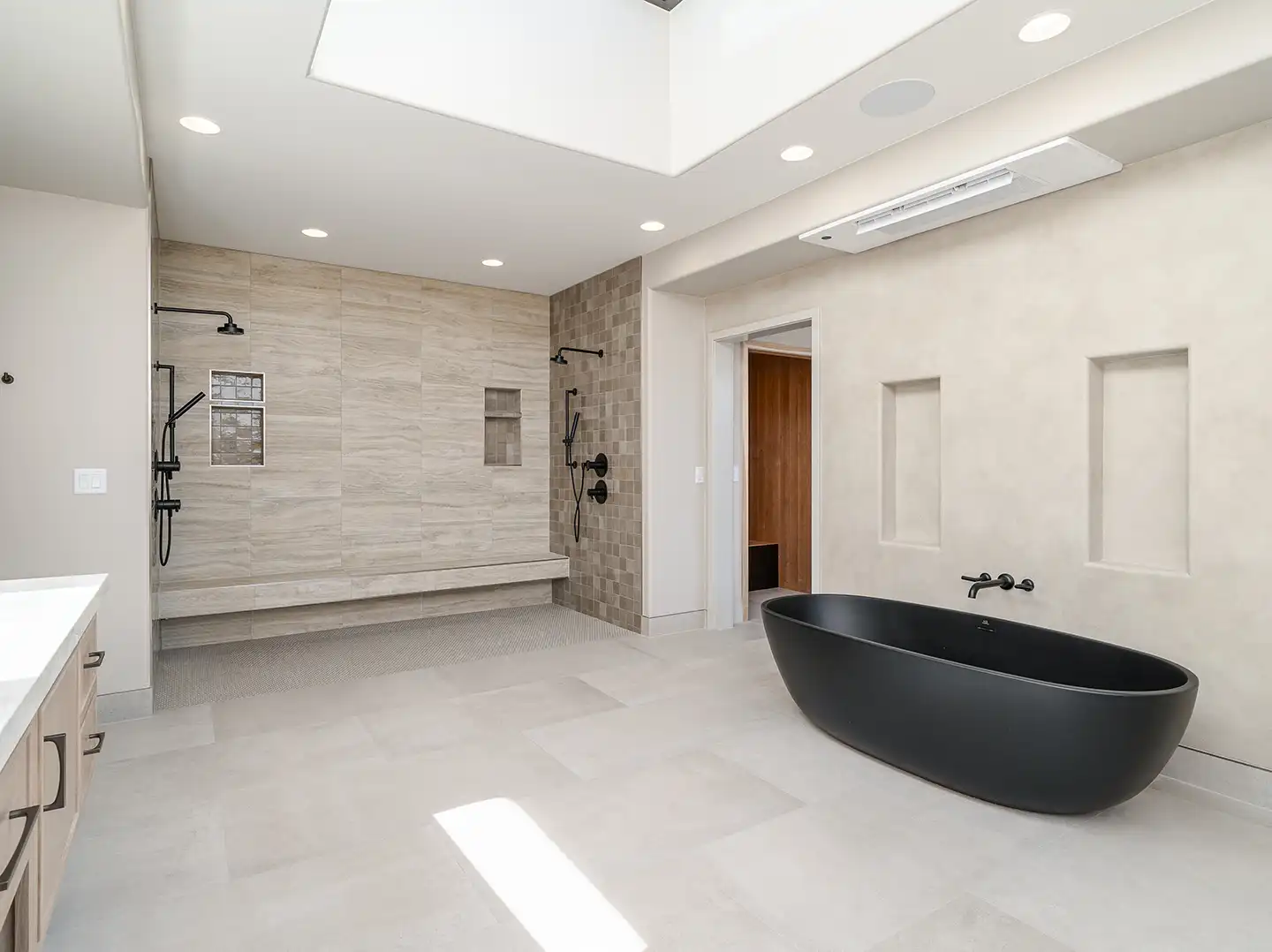 Luxury primary bathroom with black freestanding tub, skylight, dual rain showerheads, and natural stone tile walls.