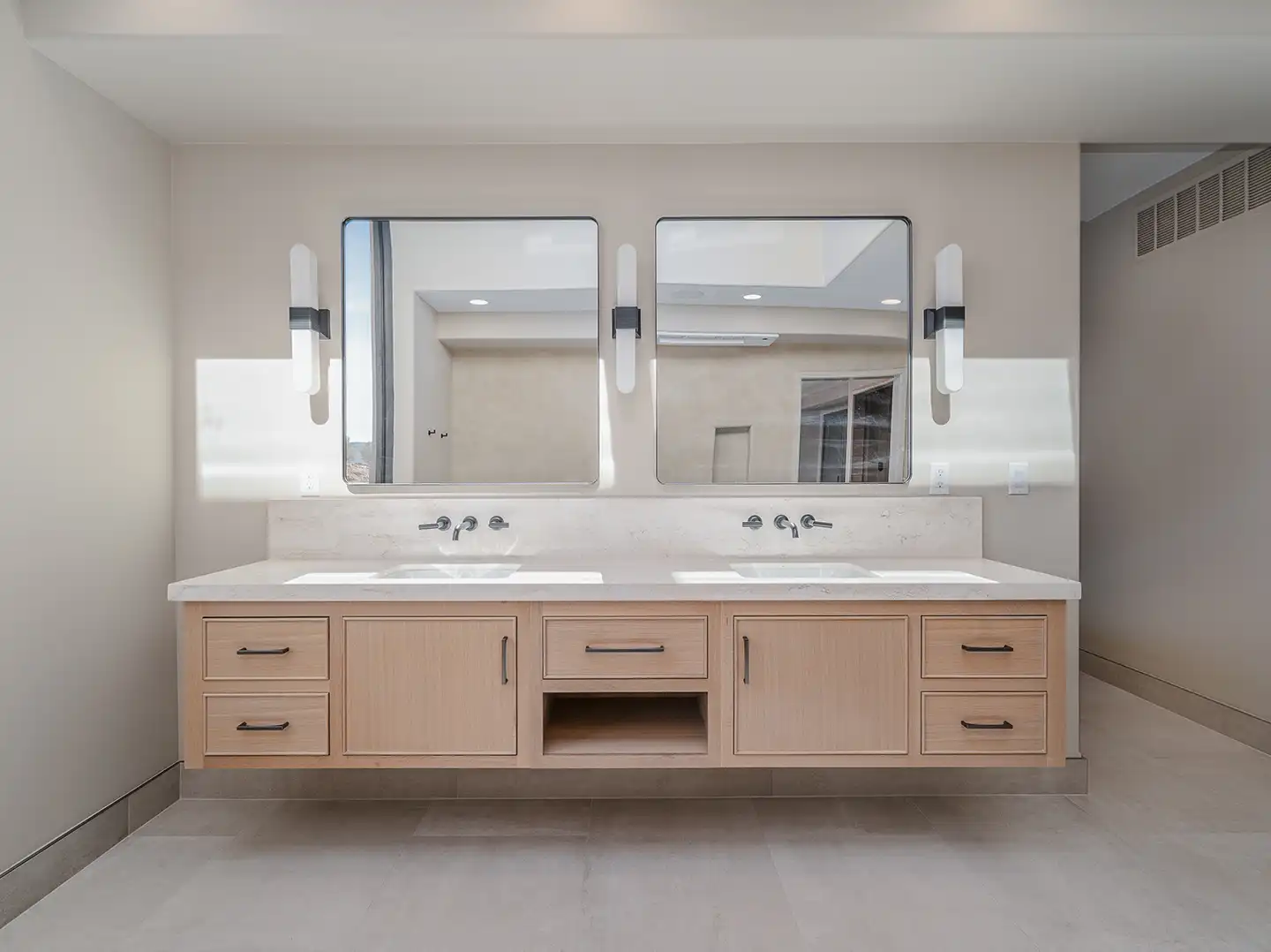 Double vanity with light oak cabinetry, quartz countertop, wall-mounted faucets, and twin rounded-edge mirrors with sconce lighting.