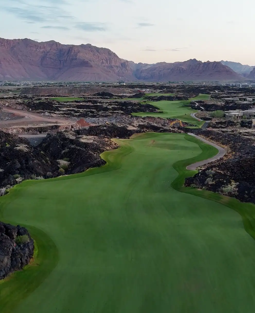 Aerial view of a winding green fairway carved through black volcanic lava rock with red mountains at sunset
