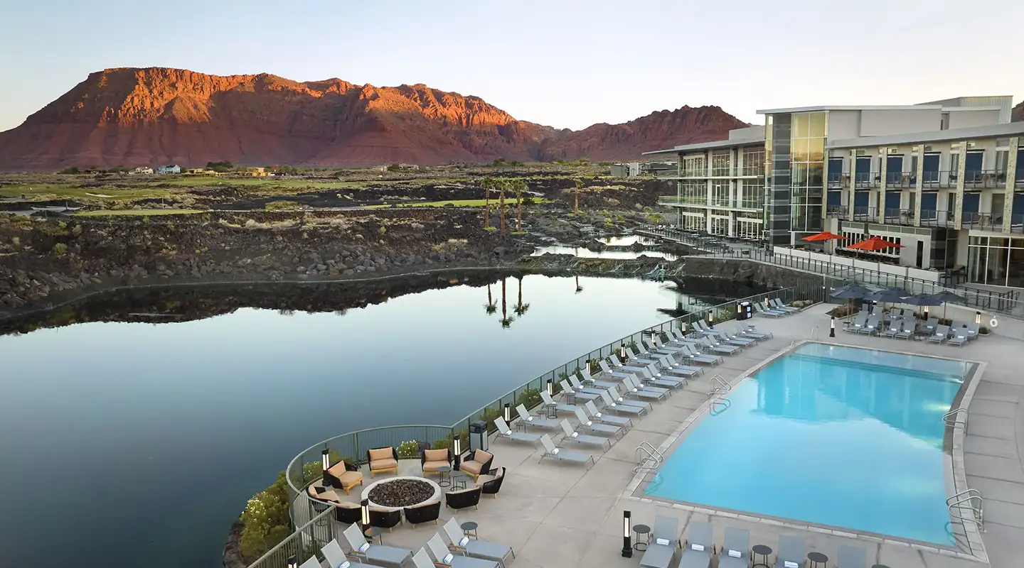 Resort pool and lounge deck beside a calm lake reflecting a red sandstone mountain at golden hour