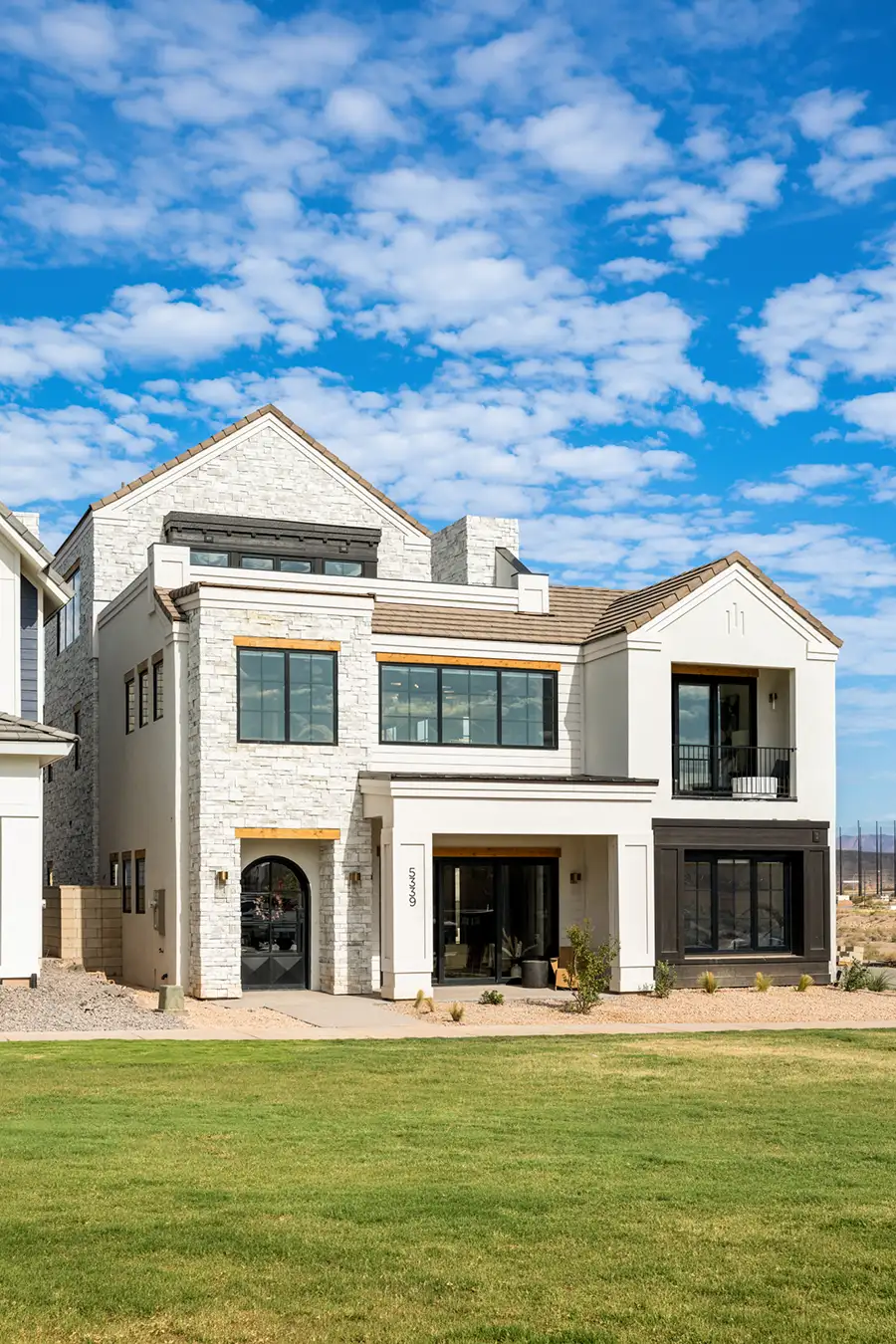 Two-story white stone-clad home with dark-framed windows, a covered entry, and a manicured green lawn under blue sky