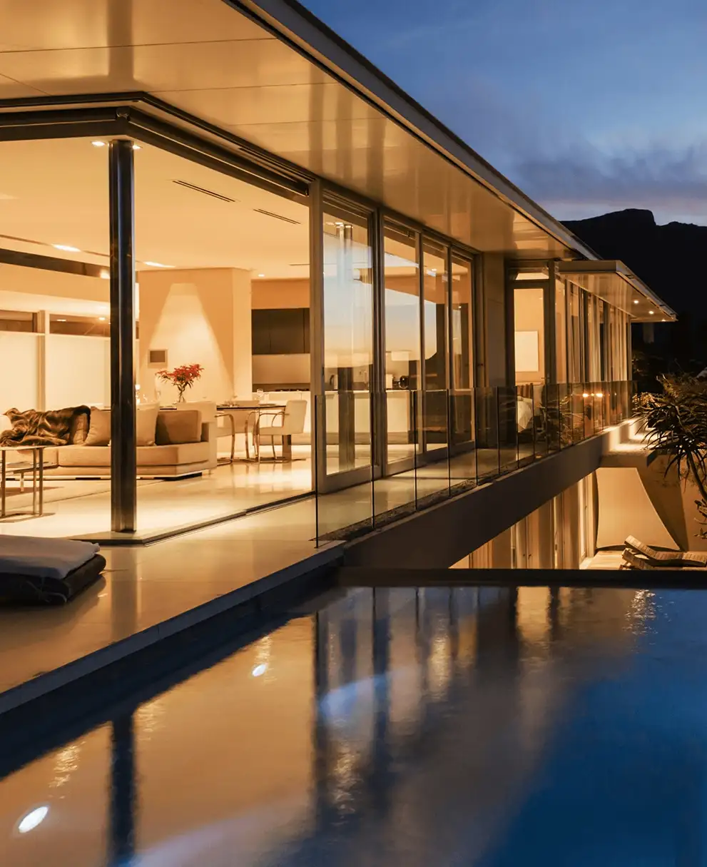 Modern luxury home with floor-to-ceiling glass walls and infinity pool reflecting warm interior lighting at twilight