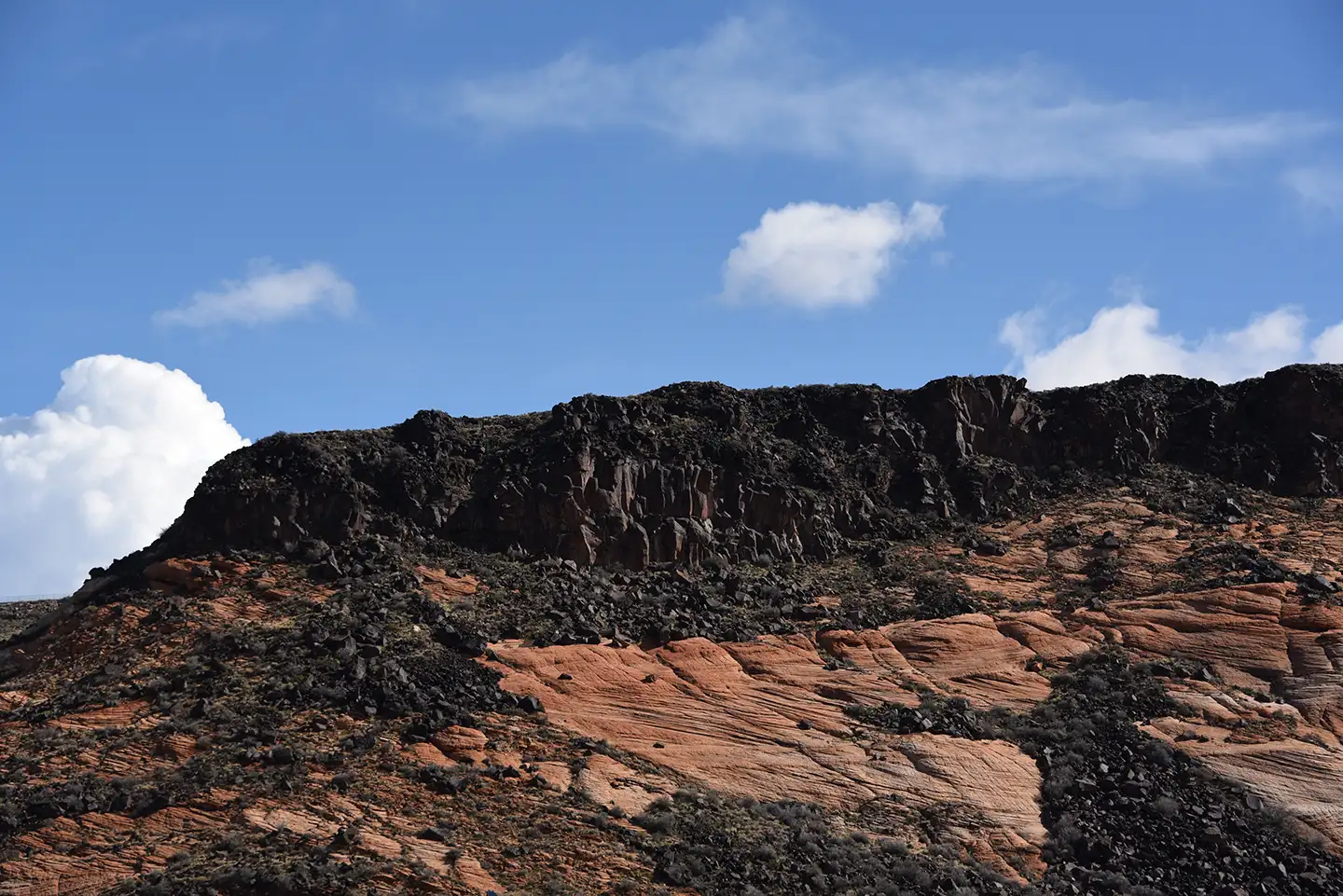 Red sandstone hillside topped with dark volcanic basalt rock under a bright blue sky with white clouds