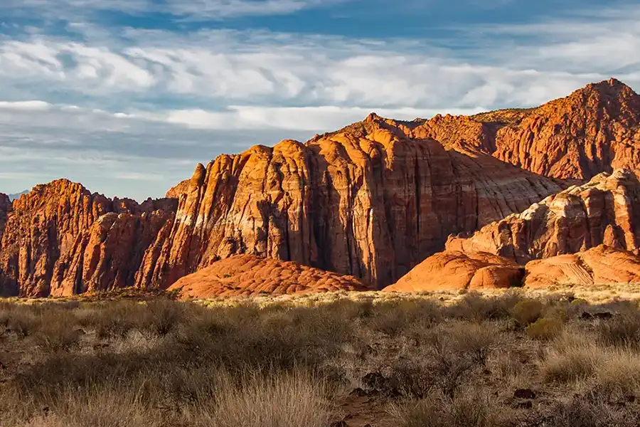 Dramatic red rock sandstone cliffs rising above desert brush under a blue sky with wispy clouds
