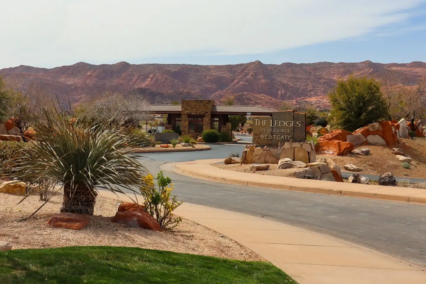 Gated community entrance with stone sign, landscaped driveway, palm trees, and red mountains behind