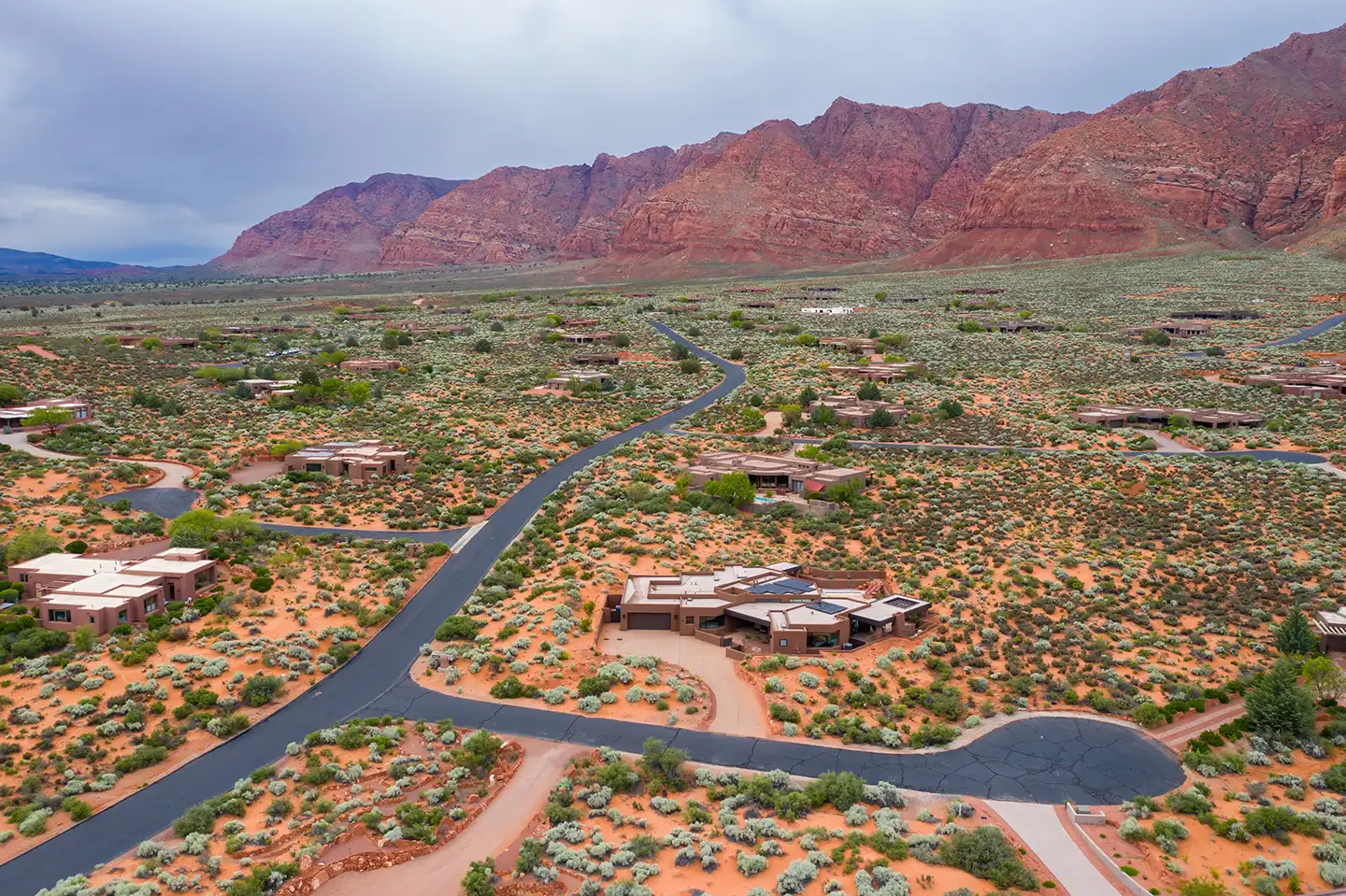 Aerial view of luxury desert homes on red sand lots with a winding road leading toward red rock cliffs
