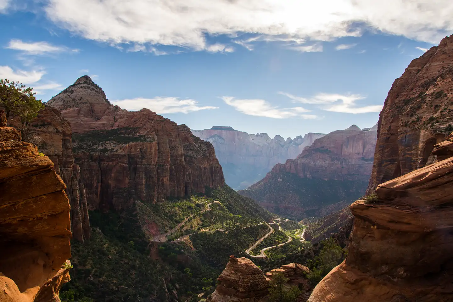 Dramatic canyon overlook framed by red rock walls with a winding switchback road through a lush green valley