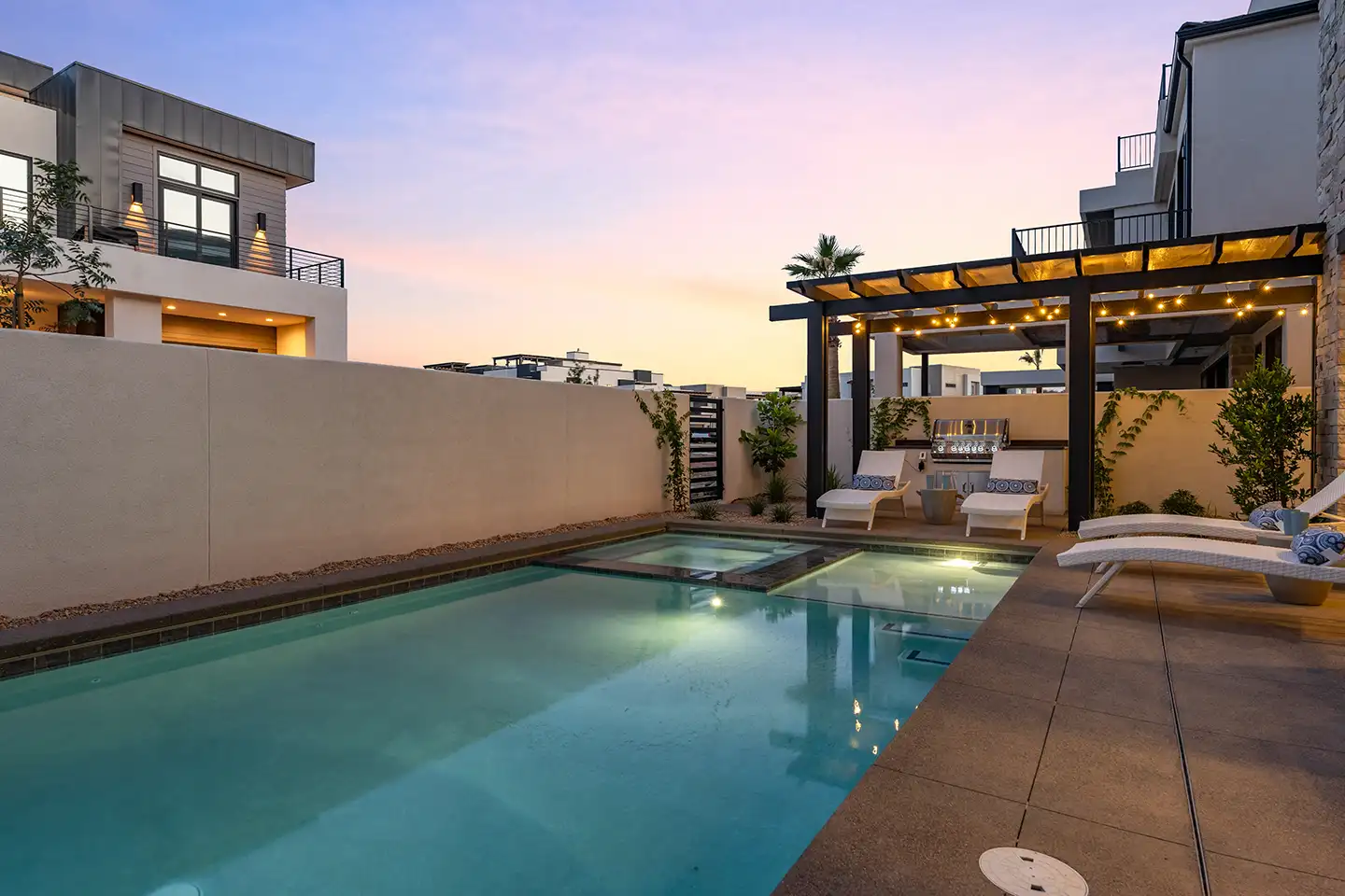 Private pool and spa with pergola, string lights, and lounge seating between modern desert homes at dusk
