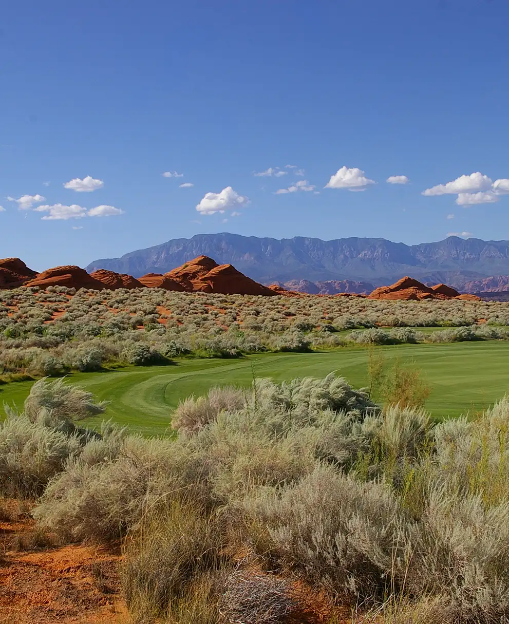 Desert golf course fairway surrounded by red sandstone formations with mountains and blue sky in the background