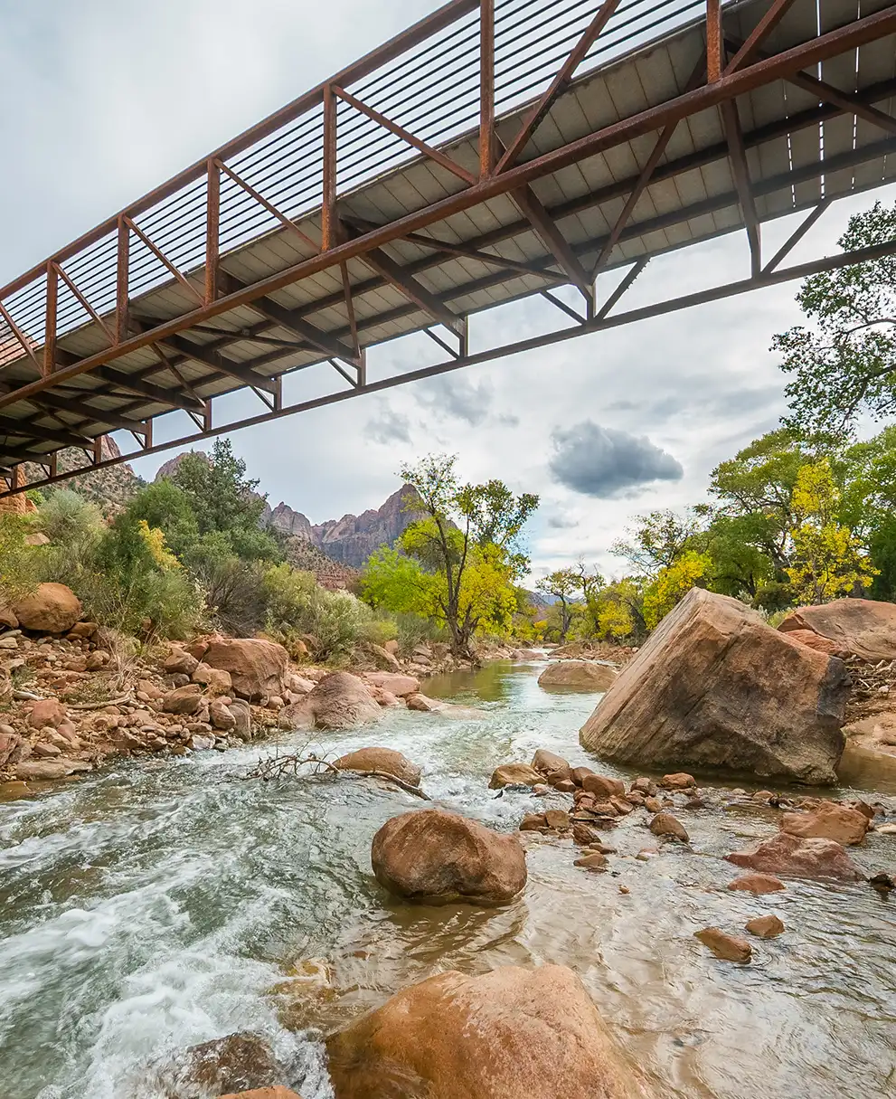 River flowing over rocks beneath a metal pedestrian bridge with red rock cliffs and lush trees in the background