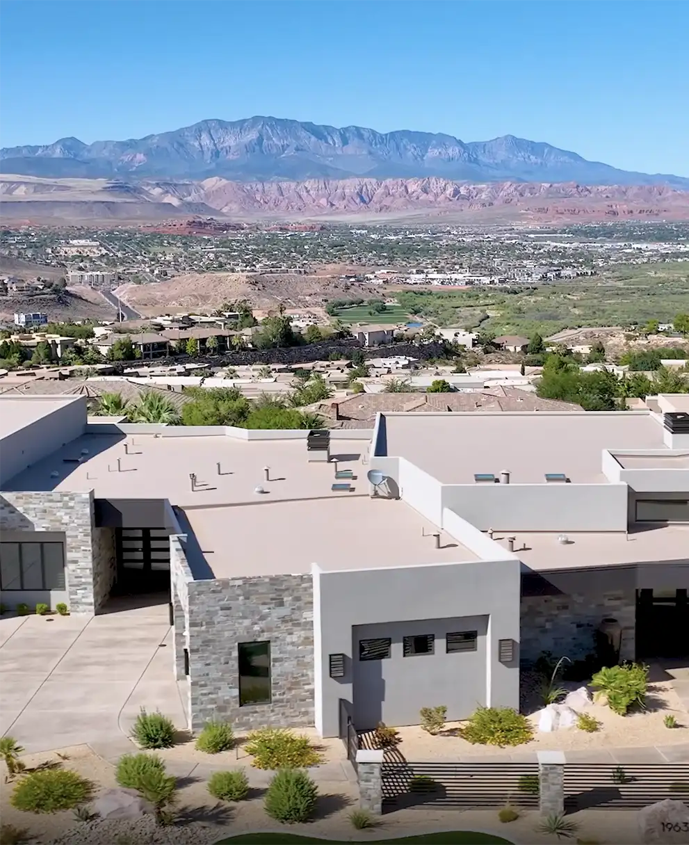 Aerial view of a modern white and stone-accented home with flat rooflines against red rock cliffs and mountains