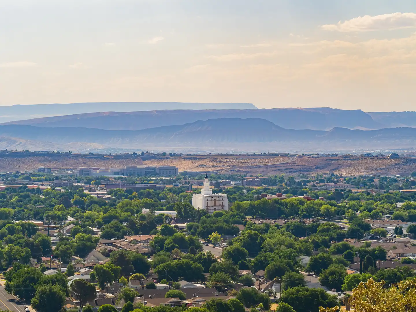 Elevated view of a tree-lined valley town with a prominent white temple building and hazy mesa cliffs beyond