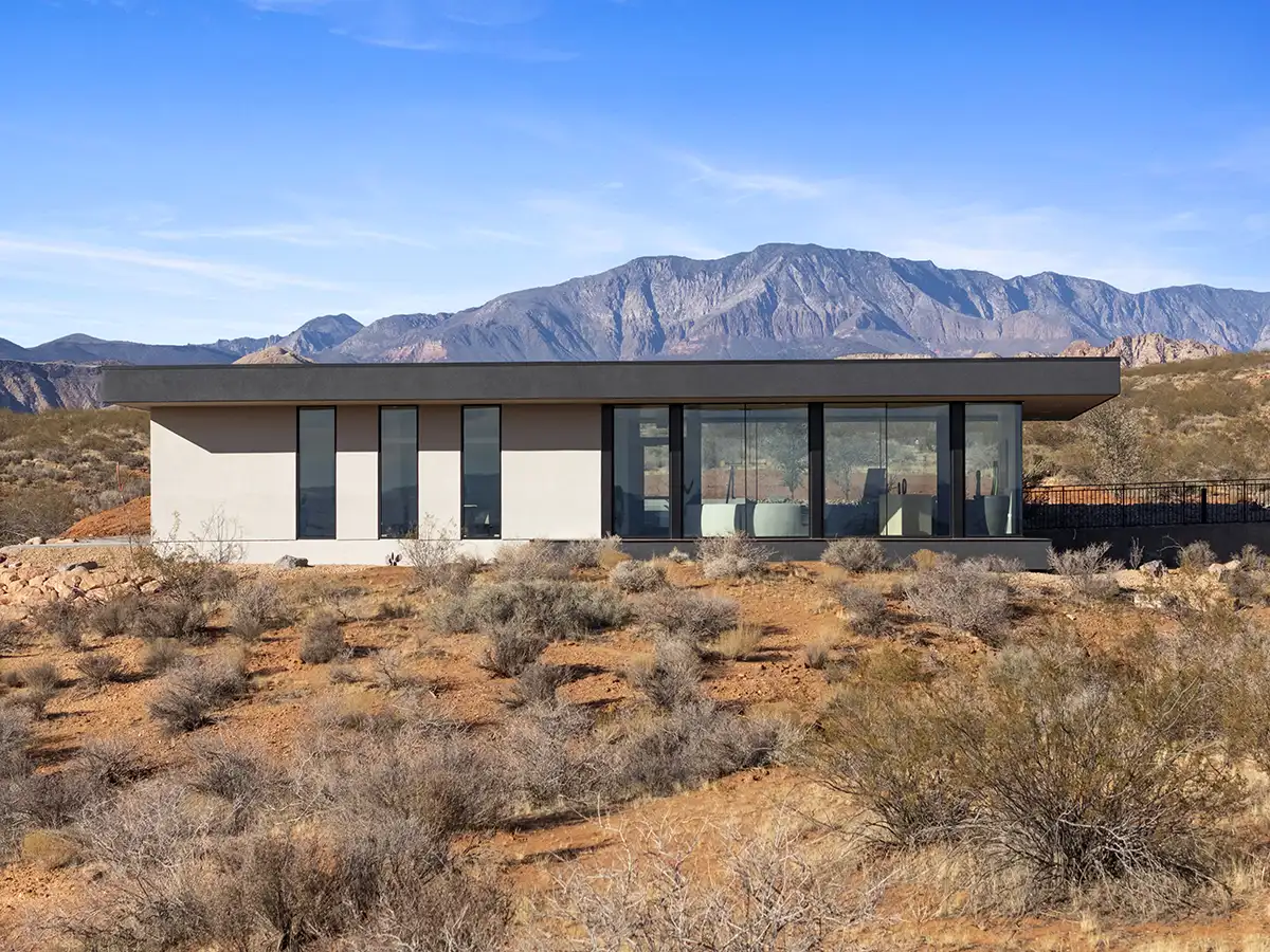 Minimalist modern home with expansive glass walls nestled in desert brush with dramatic mountain peaks rising in the background.