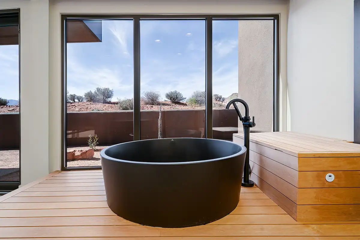 Round black freestanding soaking tub on a natural wood deck with floor-to-ceiling windows overlooking a red rock desert landscape.