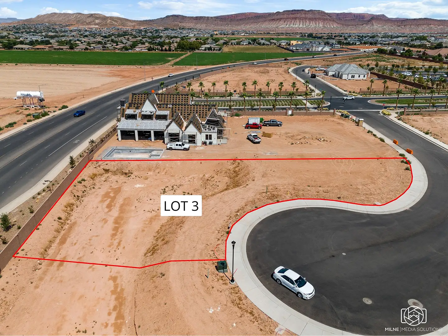 Aerial view of a home lot looking east with adjacent new construction, paved roads, and red desert mountains in the distance