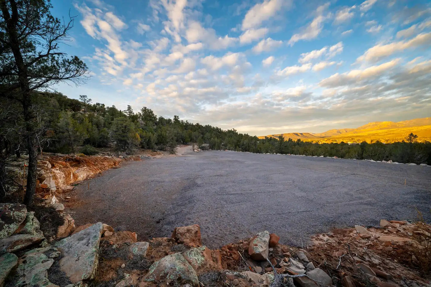 Cleared building lot at sunset with broken rocks in the foreground and tree-lined hills beneath glowing clouds.