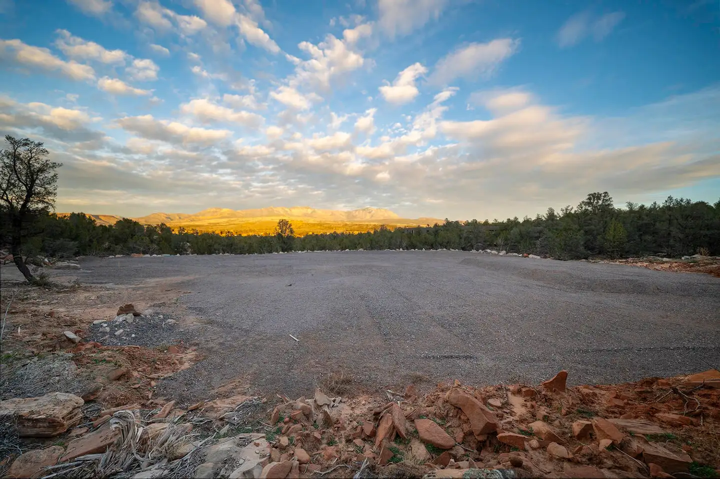 Wide cleared homesite viewed from a rocky outcrop, with pines, distant mountains and golden hills under a blue sky.