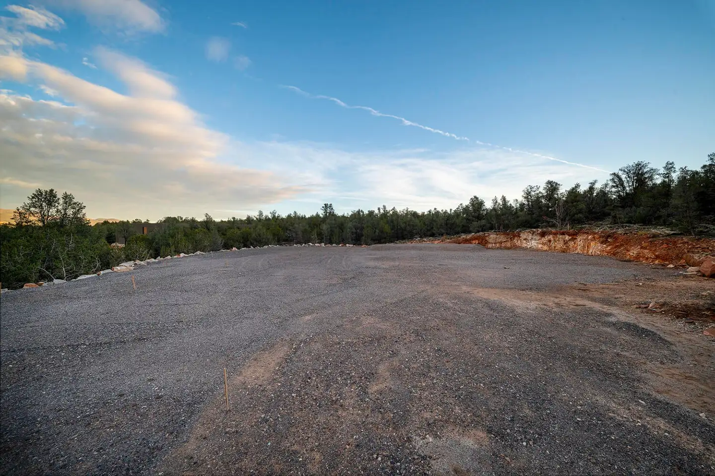 Wide cleared dirt lot under a blue evening sky, ringed by evergreen trees and red-orange exposed soil.