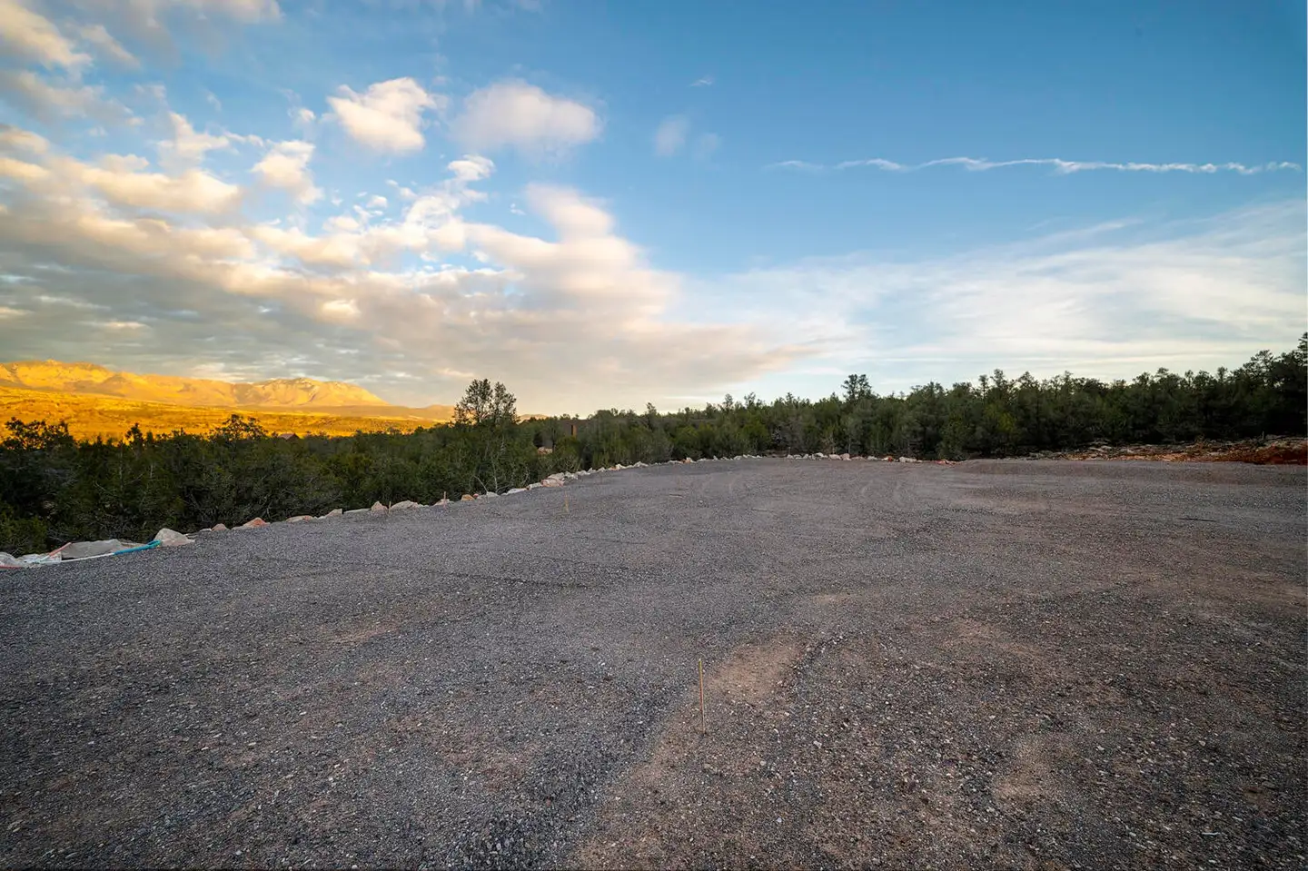 Cleared gravel building lot at sunset with golden clouds, distant pine-covered hills and rolling terrain on the horizon.