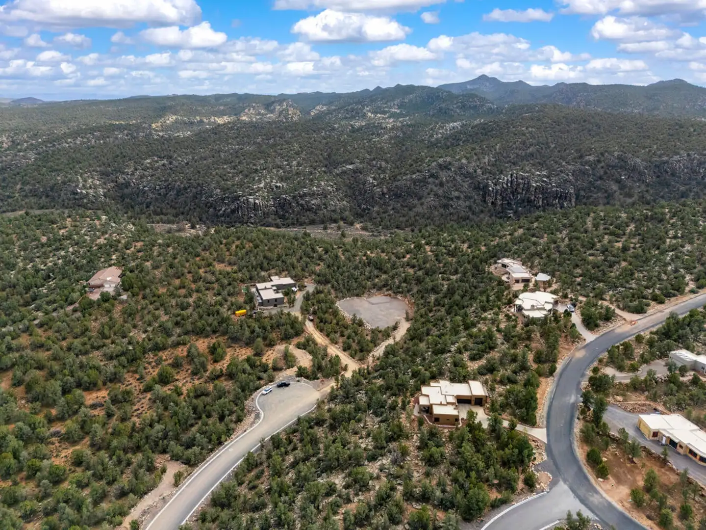 Aerial drone view of homes set into juniper-covered hills, with rocky cliffs and forested ridges behind them.
