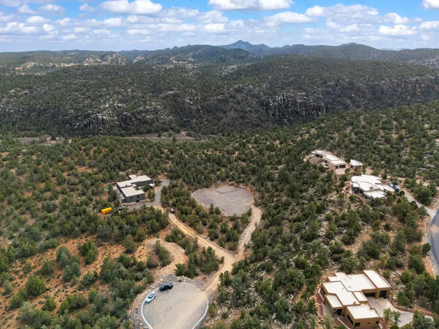 Aerial of a hillside neighborhood with paved drives, surrounded by pinyon-juniper forest and distant rocky cliffs.