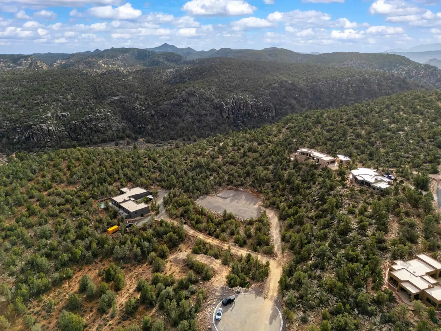 Aerial drone view of homes perched along the edge of a forested canyon under a partly cloudy sky.