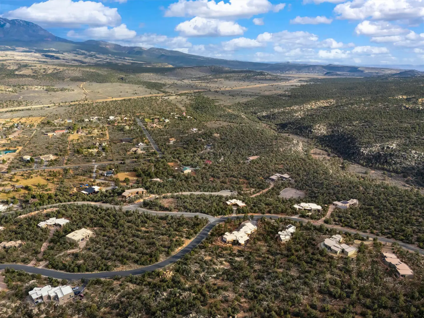 Aerial of scattered homes across a pinyon-juniper valley with rolling hills and distant mountains under puffy clouds.