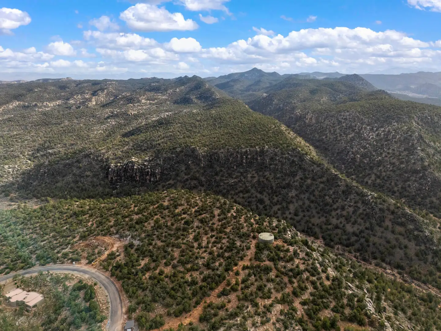 Aerial of forested mountains and rocky ridges with a small white water tank visible on a hillside in the foreground.