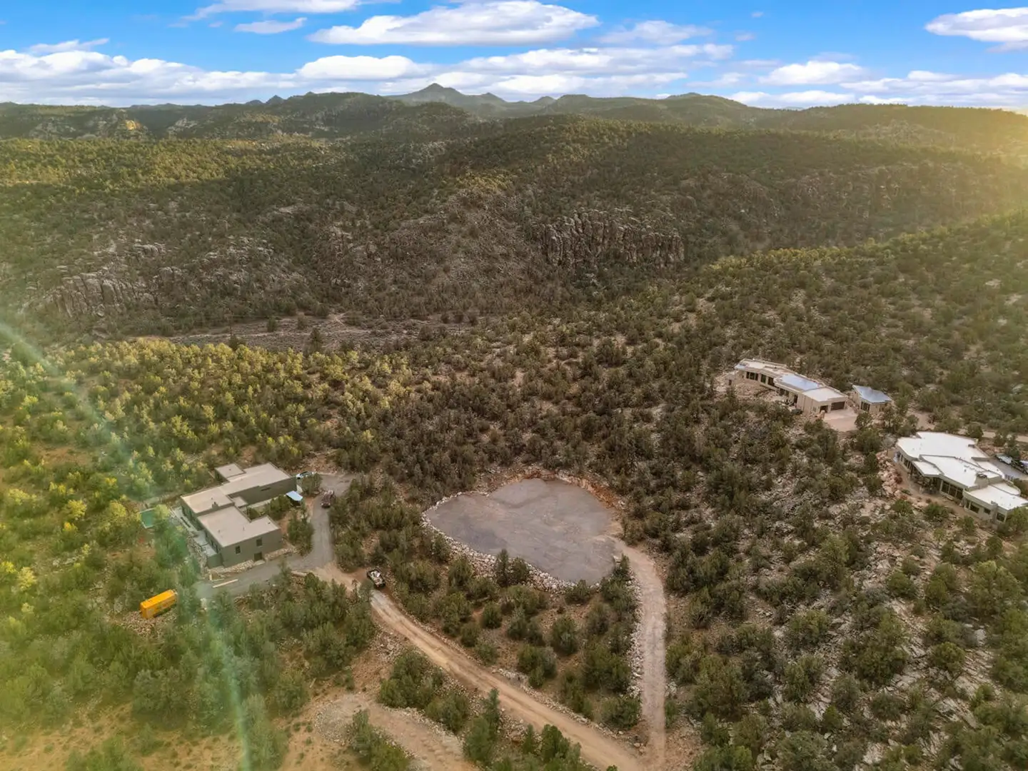 Aerial of a cleared homesite among forested high-desert hills, with warm sun flare across the right side of the frame.