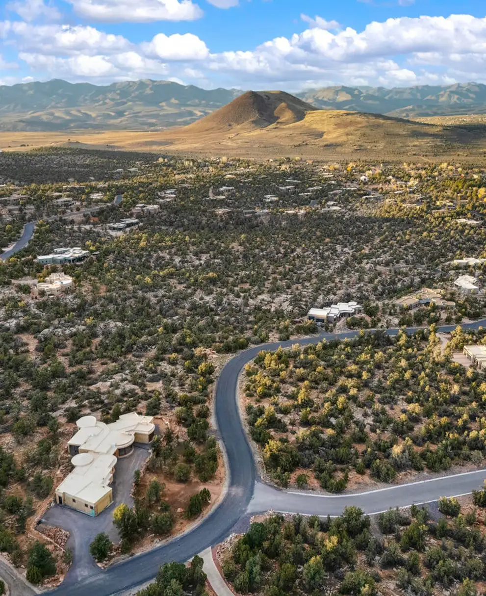 Aerial view of a curving road through a wooded high-desert neighborhood with a small cone-shaped hill and mountain ridges behind.