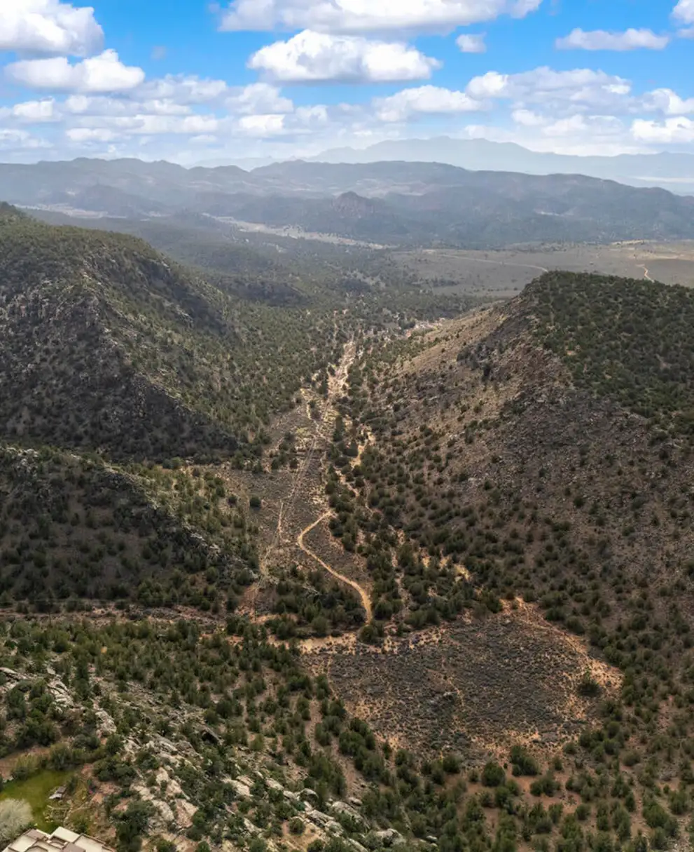 Aerial view of a forested canyon and dry wash between juniper-covered hills, with hazy mountain ranges on the distant horizon.