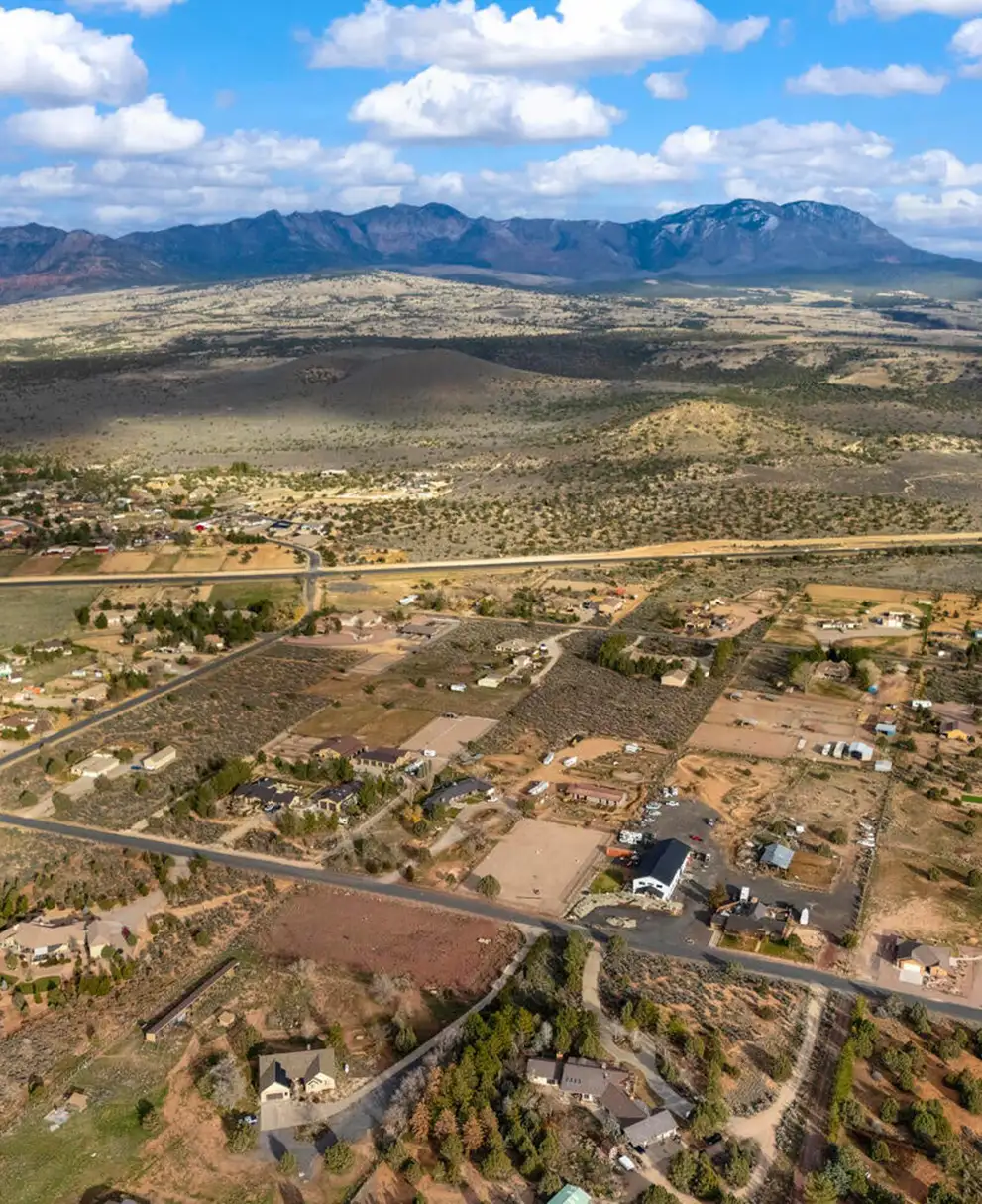 Aerial view of a rural high-desert subdivision with scattered homes, fields and a paved road below a long mountain range.