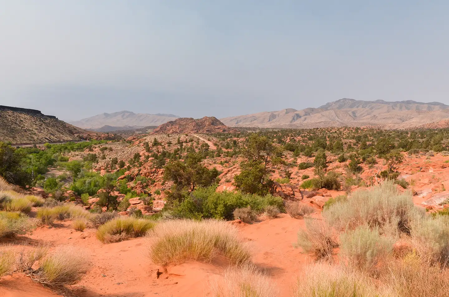 Open Southern Utah desert with red sand, scattered shrubs, and rolling mountain ridges fading into the haze.