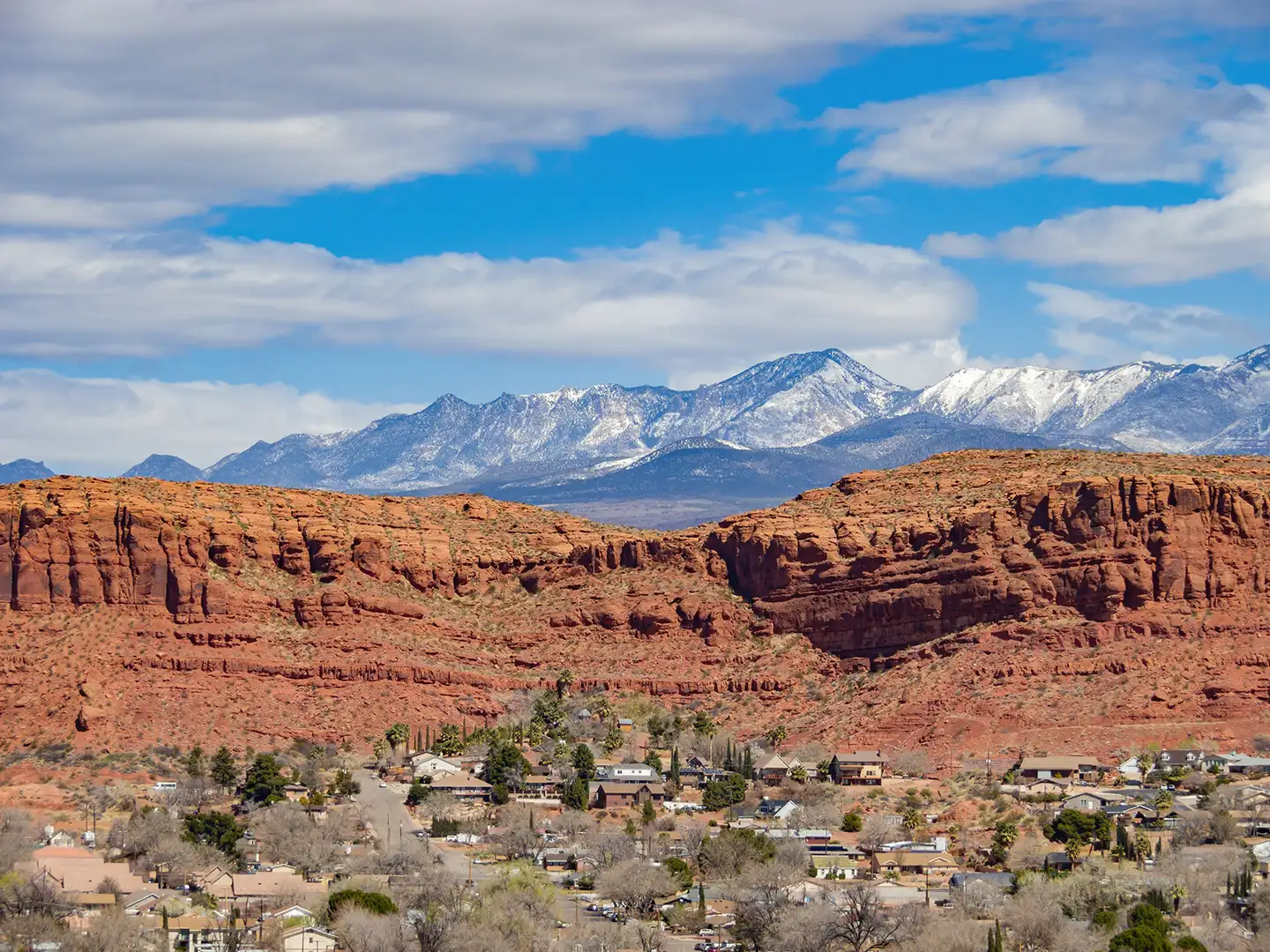 Red rock cliffs rise above a Southern Utah neighborhood, with snow-capped mountain peaks beyond and homes nestled at the base of the bluffs.