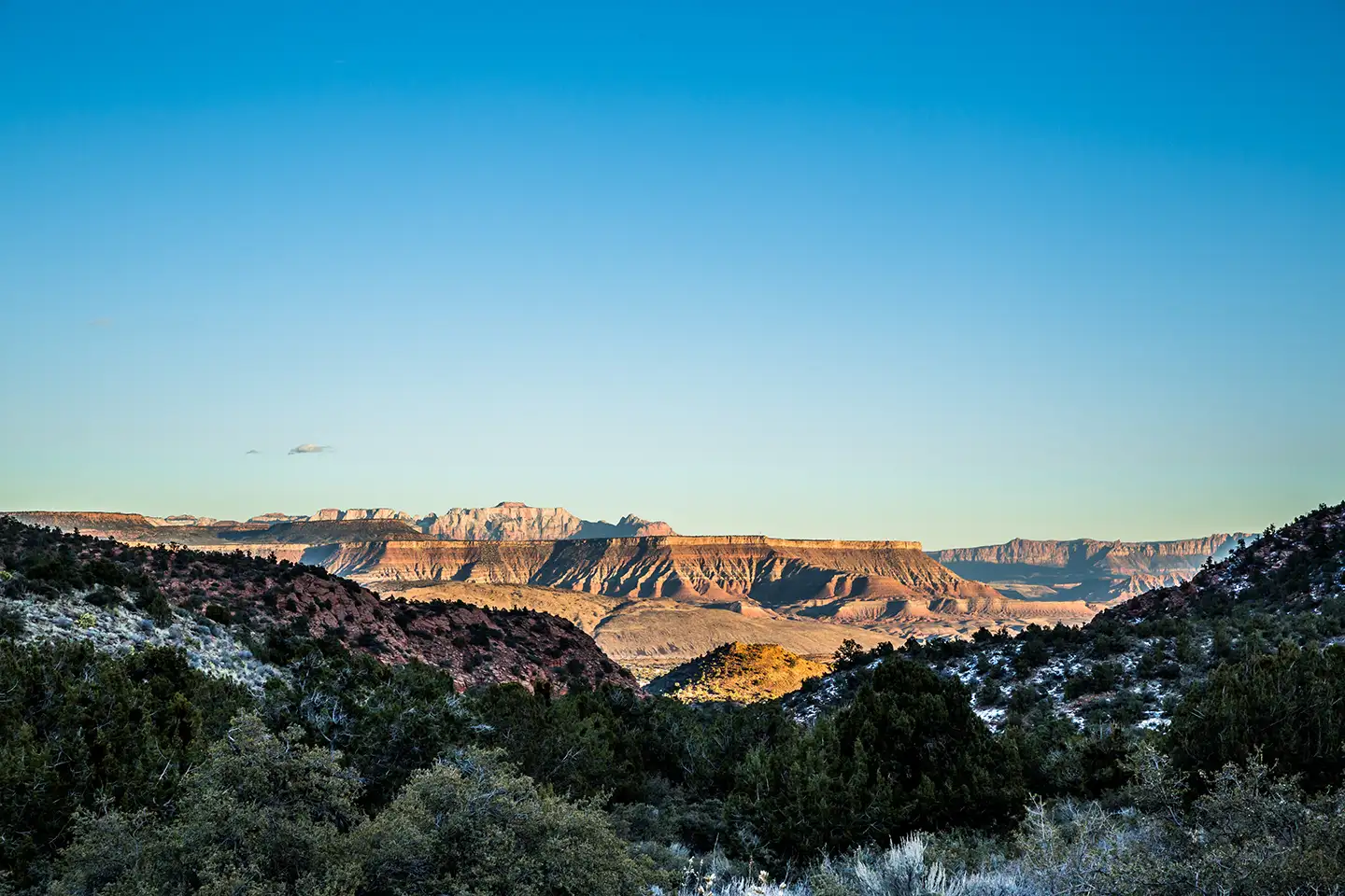 A wide overlook of green pinyon-juniper terrain near Pine Valley, with sandstone mesas catching warm light along the horizon under a clear sky.