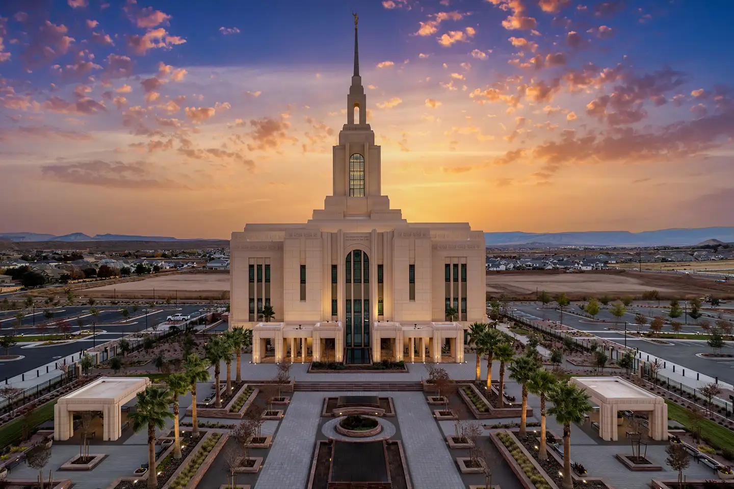 The Red Cliffs Utah Temple lit at dusk, with a glowing pastel sky behind the spire and palm-lined gardens at the base.