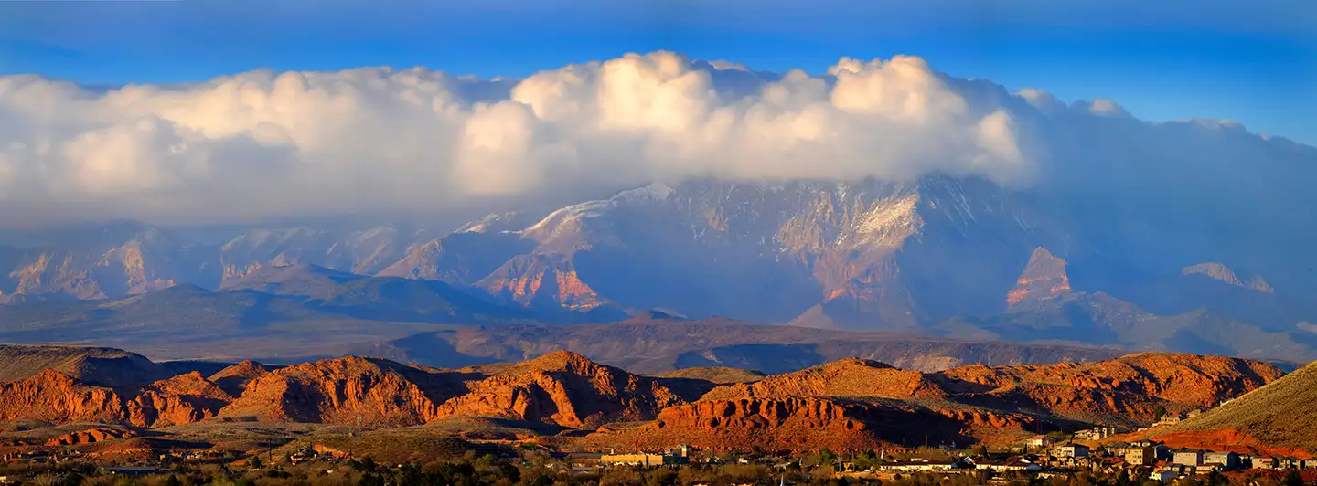 Snow-capped Pine Valley Mountain rises behind red sandstone hills above St. George, with thick clouds clinging to the peaks.