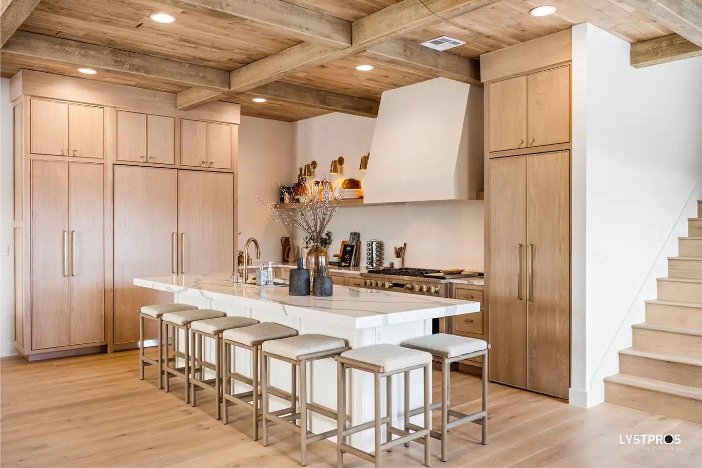 Open-plan luxury kitchen with light oak cabinetry, exposed wood beam ceiling, a marble waterfall island, and brass pendant detailing.