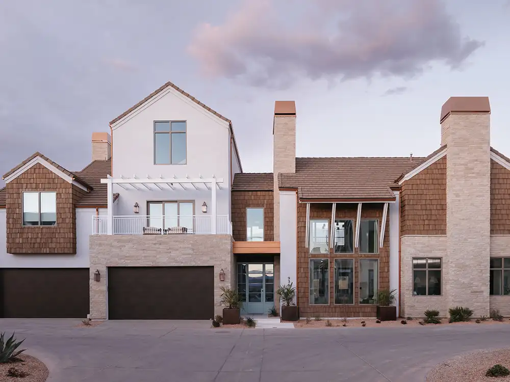 Coastal craftsman home with cedar shake siding, double stone chimneys and second-story balcony at dusk.