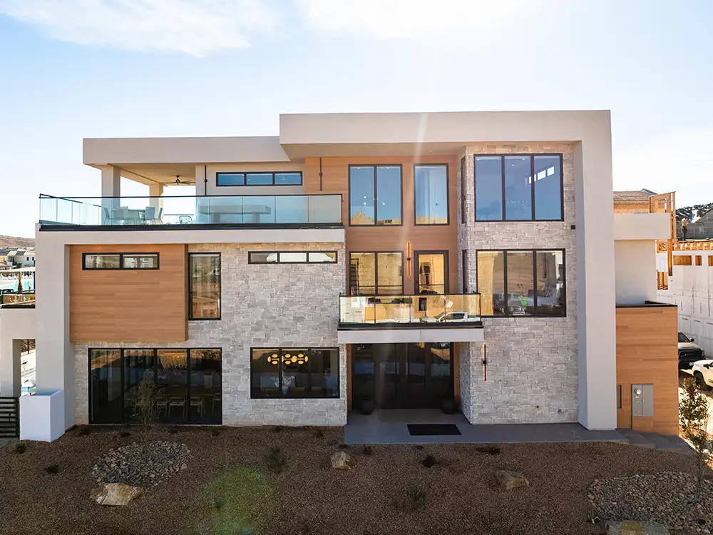 Three-story modern home with stone, wood and glass-railed balcony backed by desert hills at dusk.