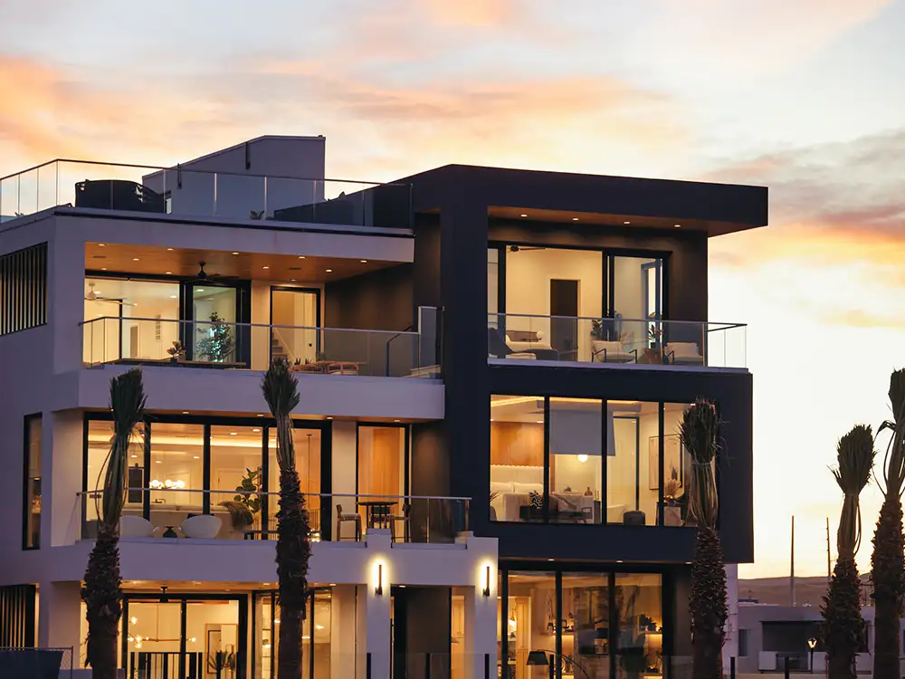 Modern multi-story home at sunset with glass balconies, palm trees and warm interior lights glowing through large windows.