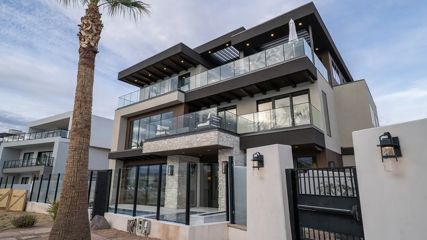 Modern three-story home exterior with stone accents, glass balconies, a tall palm tree, and a dark gated entry.