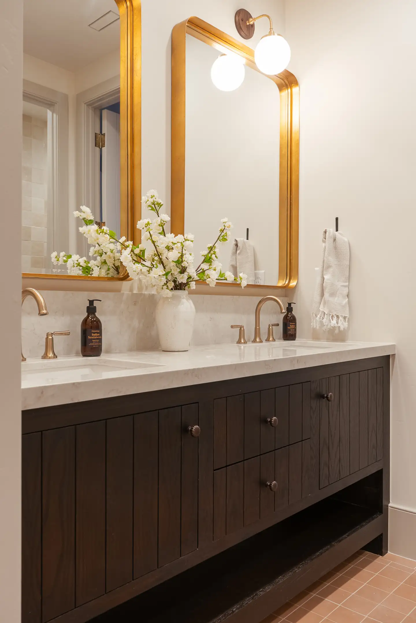 Bathroom double vanity with dark wood cabinetry, brass-framed mirrors, white blossoms in a vase, and brass faucets.