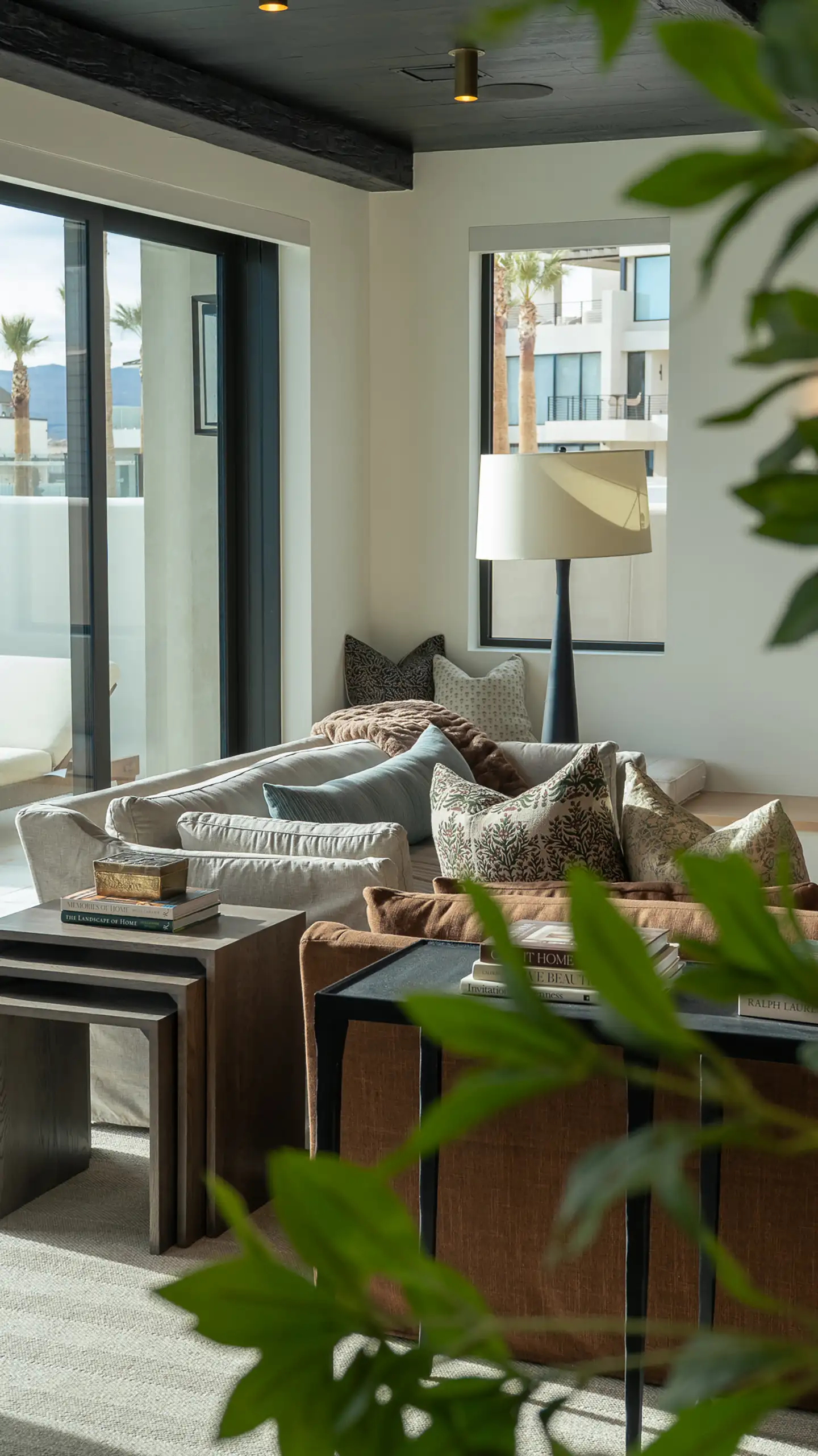 Living room corner with a brass-trimmed floor lamp, brown sofa with patterned pillows, and houseplant leaves in the foreground.