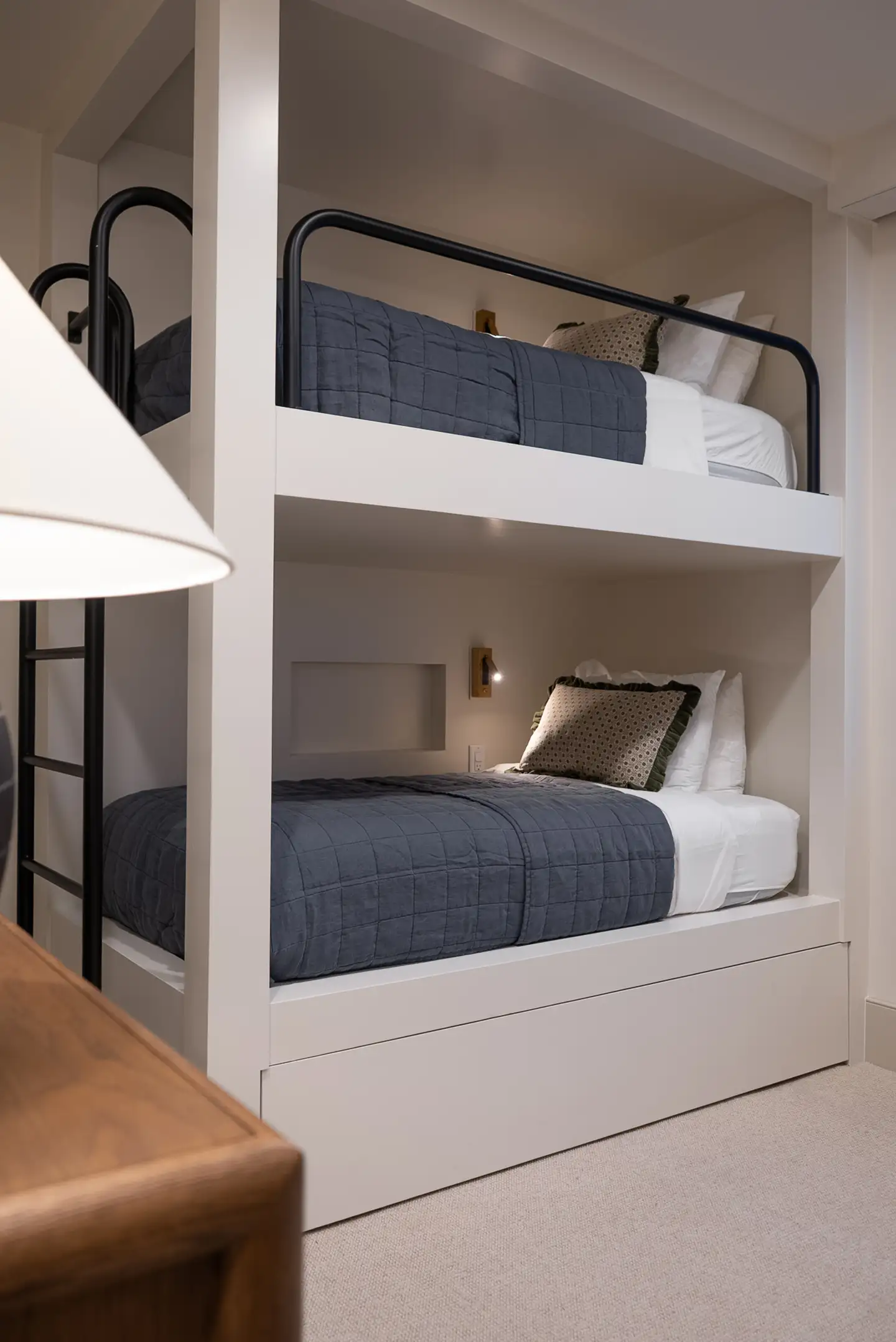White built-in bunk beds with navy quilted bedding, decorative pillows, and a black ladder for upper bunk access.