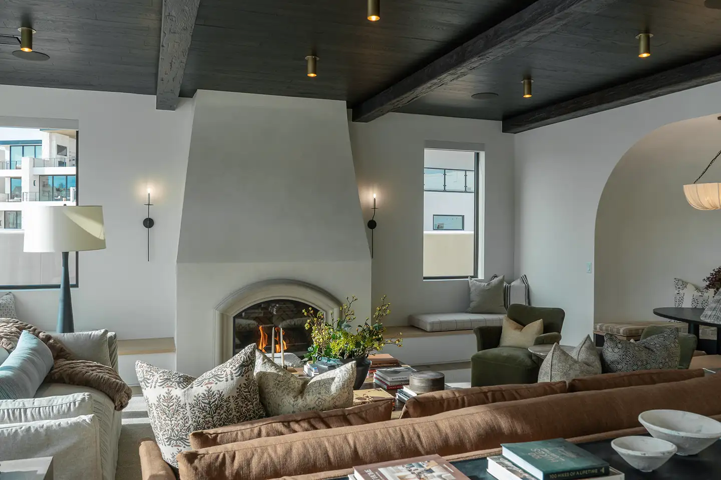 Living room with a tall white plaster fireplace, dark beamed ceiling, brown sectional, and a built-in cushioned window seat.