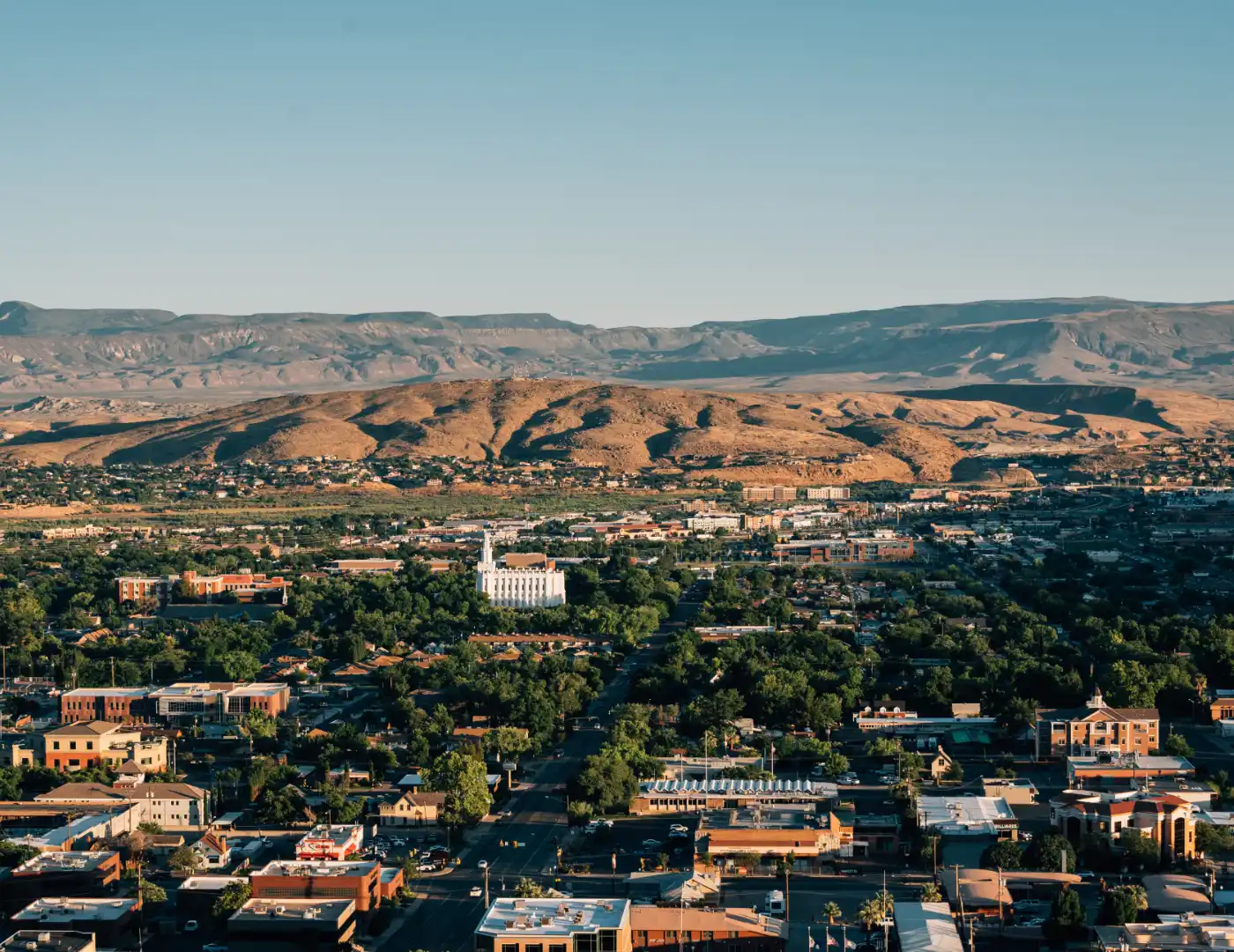 Hilltop view of a downtown skyline with a tall white temple, leafy streets, and a rugged mountain ridge in the distance under clear sky